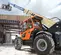 Side view of an orange and yellow JLG telehandler with extended boom, forklift attachment and cabin on a construction site.